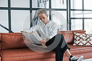 Reads newspaper. Caucasian young guy in elegant white shirt indoors at home