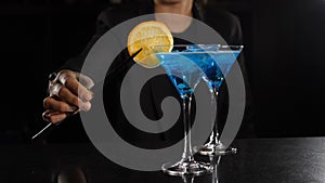 Ready cocktail is on the bar at nightclub. Female bartender decorating cocktail with blue liquid with slice of lemon