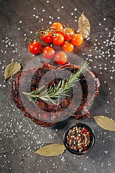 Ready meat steak on a dark metal cutting board, next to a sprig of aromatic rosemary, tomatoes, a black mug with mixed peppercorns