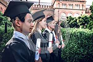 Ready for new beginnings!Happy graduates are standing in a row in university outdoors in mantles with diplomas in hand