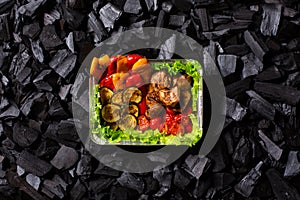 Ready shish kebab. Portion of grilled meat and vegetables in a disposable container on charcoal background. Copy space.