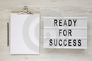 `Ready for success` words on a lightbox, clipboard with blank sheet of paper on a white wooden background, top view. Overhead,