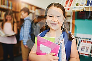 Ready to do some research now. Portrait of a cheerful young girl holding school books while standing inside of a library