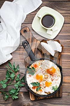 Ready-to-eat breakfast: shakshuka from fried eggs with tomatoes and parsley in a pan, bread with butter and coffeee