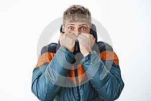 Ready for winter. Scared good-looking european blond man with blue eyes getting warm pulling collar of puffer jacket on