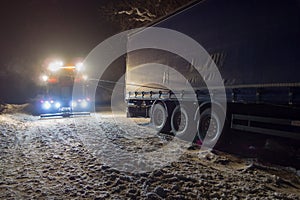 Truck traffic accident at night, on a snowy winter road. Strongly illuminated Wrecker truck pulls a truck out of snow hanging.