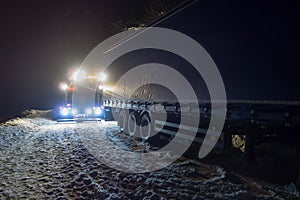 Truck traffic accident at night, on a snowy winter road. Strongly illuminated Wrecker truck pulls a truck out of snow hanging.