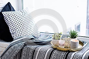 Real apartment interior decor, aromatic candles and plants on vintage tray with pillows and blanket on white windowsill