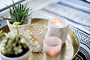 Real apartment interior decor, aromatic candles and plants on vintage tray with pillows and blanket on white windowsill