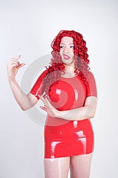 Real doll woman wearing red latex rubber dress and posing on white studio background alone. curvy plus size adult girl standing as