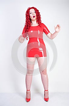 Real doll woman wearing red latex rubber dress and posing on white studio background alone. curvy plus size adult girl standing as