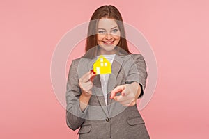 Real estate agency. Beautiful positive woman in elegant business suit holding paper house and pointing to you