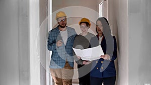 A real estate agent showing a new draft apartment to a young married couple in helmets holding a layout on the paper