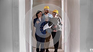 A real estate agent showing a new draft apartment to a young married couple in yellow helmets holding a layout on the