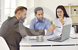 Real estate agent sitting at desk, talking to young couple and showing them something on laptop