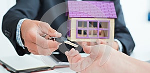 Real estate agent in suit holds house model in one hand and in the second hand holds out the keys of the house to woman.