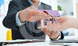 Real estate agent in suit holds house model in one hand and in the second hand holds out the keys of the house to woman.