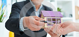 Real estate agent in suit holds house model in one hand and in the second hand holds out the keys of the house to woman.