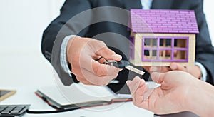 Real estate agent in suit holds house model in one hand and in the second hand holds out the keys of the house to woman.