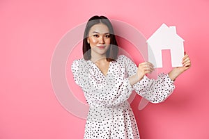 Real estate. Beautiful asian woman demonstrating paper house model, looking at camera confident, advertising home for