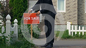 Real estate broker in suit setting for sale sign front of house, agency service