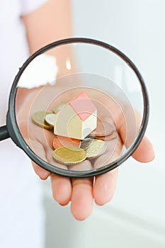 Real estate concept - coins and house architectural model in woman hand under magnifying glass