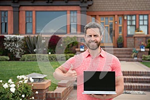 Real estate manager. Happy businessman standing outside near his new home. Suburban house. Young man holding laptop