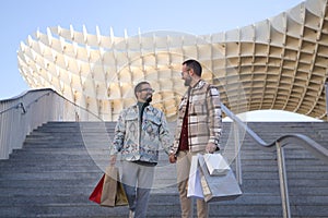 Real marriage of gay couple, holding hands, walking down some stairs and with many shopping bags in the other hand, happy and