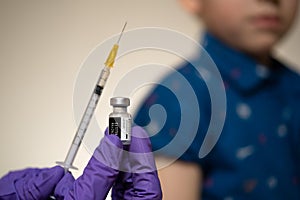 Real Pfizer-BioNTech vaccine vial hold in hand with syringe and child sitting with rolled up sleeve on the blurred background. Edi