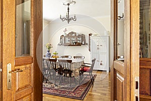 Real photo of an antique dining room interior with a big table, chairs, wall cupboard and carpet. View through a door