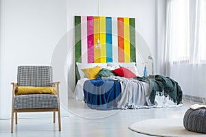 Real photo of a bright bedroom interior with white walls, comfy armchair and colorful blankets on a double bed