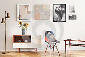 Real photo of bright eclectic living room interior with many posters, colorful chair, wooden cupboard with flowers and coffee tabl photo