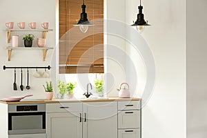 Real photo of a bright kitchen interior with wooden counter, black lamps, white cupboards and pink accents