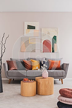 Real photo of a bright living room interior with a set of paintings hanging on a beige wall and cushions in warm colors on a couch