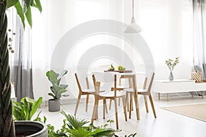 Real photo of a bright, scandi dining room with a white lamp above a round table with wooden chairs, next to the window