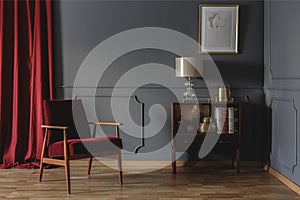 Real photo of a corner of a retro living room interior with elegant, beige lamp on a wooden cabinet next to a red armchair