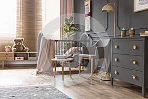 Real photo of a cot standing next to an armchair, lamp and cupboard in dark and classic baby room interior