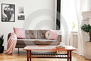 Real photo of couch with pastel pink cushions and blanket standing in white sitting room interior with wooden table with book and