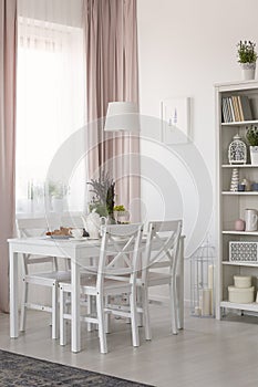 Real photo of dining room interior with chairs and white table with lavender and breakfast, window with drapes and rack with decor