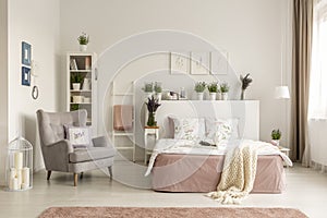 Real photo of a feminine bedroom interior with a comfy armchair, bed, plants and shelf