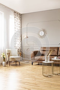 Real photo of full of light daily room with retro armchair, lamp, brown leather couch and tables