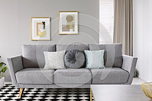 Real photo of a grey sofa with black and white pillows in a living room interior with checkered floor and posters in gold frames