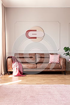 Real photo of light grey living room interior with modern clock on wall with molding, carpet on the floor and leather sofa with pi
