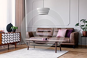 Real photo of light grey sitting room interior with window with drapes, leather sofa with pillows, coffee table on carpet and cupb