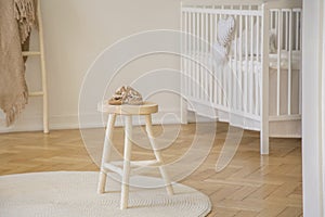 Little baby shoes placed on wooden stool standing in white kid room interior with crib, round rug and herringbone pa