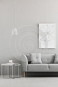 Real photo of map poster hanging on the wall in living room interior with grey sofa, metal end table with books and coffee cup and