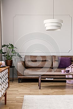 Real photo of modern lamp hanging above leather couch with purple pillow in bright living room interior with coffee table, molding