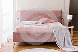 Real photo of pastel pink king-size bed with knit blanket and two pillows standing in bright bedroom interior with molding on wall