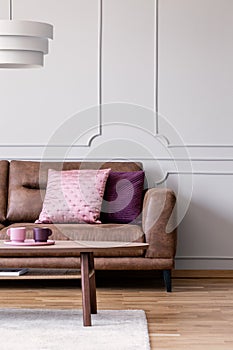 Real photo of pastel pink and violet cushions placed on leather couch in light grey living room interior with wooden coffee table