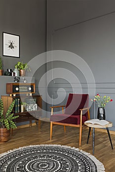 Real photo of a red armchair standing between a stool with flowers and shelf with books, radio and plants in retro living room in